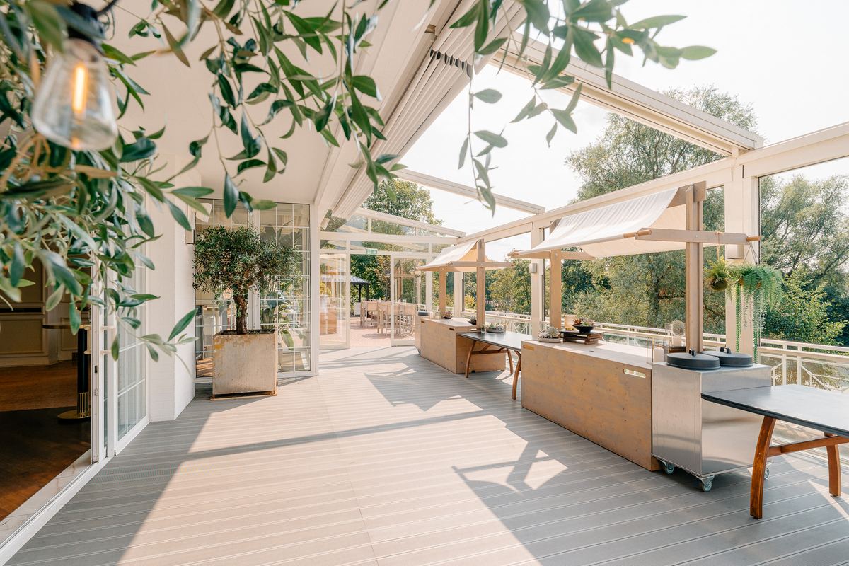 Überdachte Terrasse des Deichgraf mit Buffetstationen und Olivenbaum, im Hintergrund die Bäume des Südparks